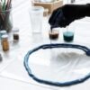 Close-up of gloved hand pouring blue resin onto a surface with art supplies.