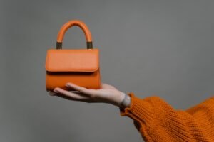 pexels photo 8801195 8801195 Stylish orange handbag held by a hand against a gray background. Perfect for modern fashion.